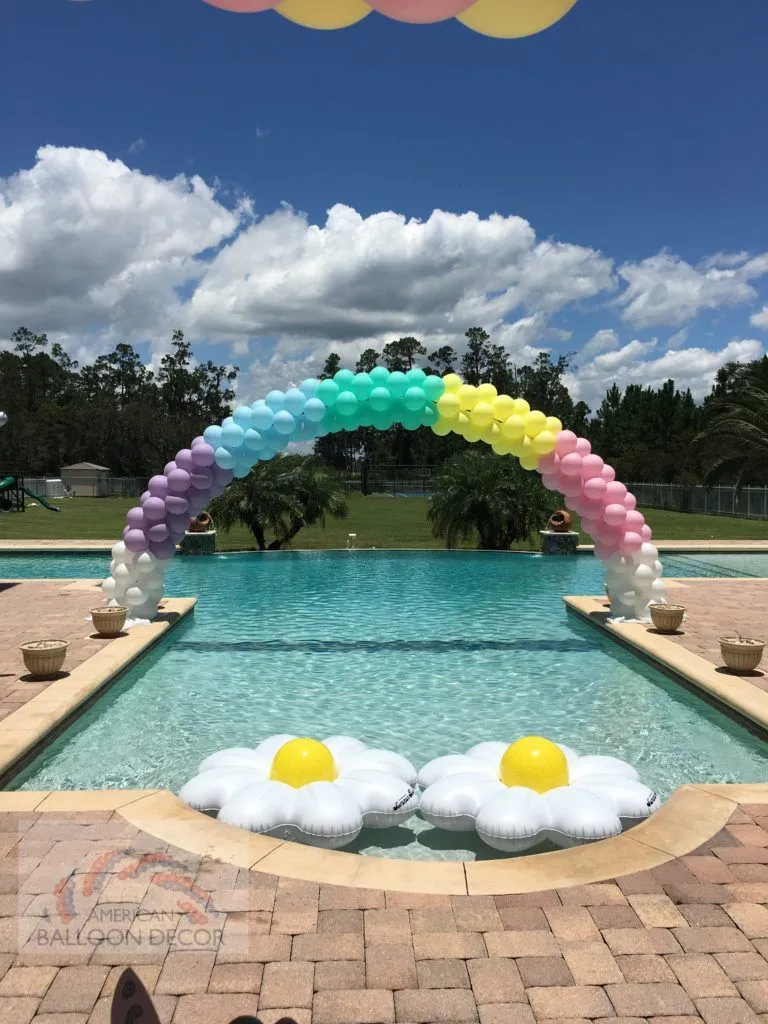 a purple, blue, yellow, and pink balloon arch that goes over a swimming pool