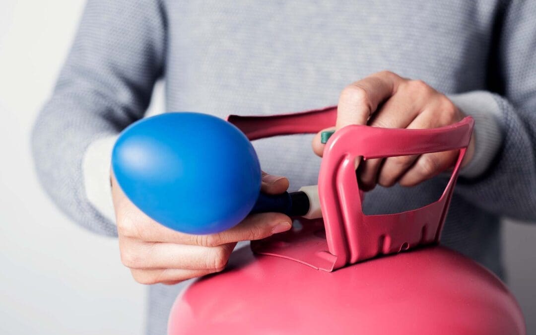 closeup of a caucasian man, wearing a gray sweater, inflating a blue balloon with helium from a pink cylinder